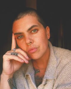 A portrait of a person with short hair and a nose ring, resting their head on their hand. They have light green eyes, tattoos partially visible on their chest, and are wearing a light-colored sweater. The image is warmly lit, creating a calm and reflective mood.